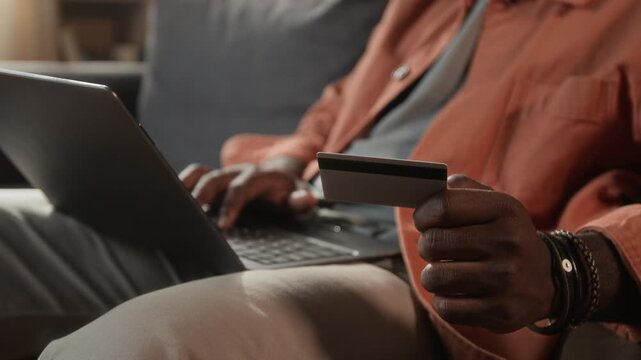 Unknown African American man holding credit card while typing on laptop keyboard during online payment process. Ideal for digital banking campaigns and secure e-commerce checkout advertisements
