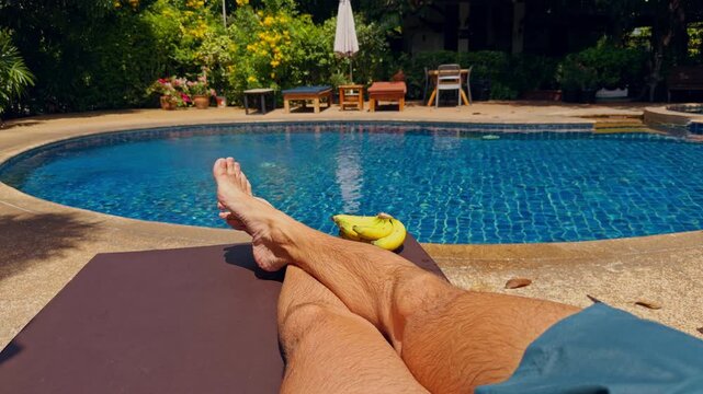 Bare Legs Rest Crossed on a Lounger Beside a Blue Swimming Pool, With Bananas and Lush Garden Greenery Framing First Person Summer Leisure and Wellbeing