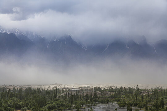 Aerial view of the Skardu valley featuring snow-capped mountains partially obscured by clouds and mist, with lush green trees and a sandy valley floor in Skardu, Pakistan.