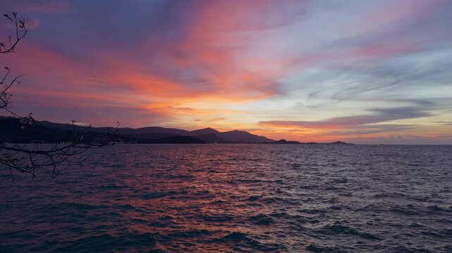 Rippling Sea Beneath a Colorful Sunset and Dark Mountain Coast, Framed by Sparse Branches, Celebrating Nature and Tranquility