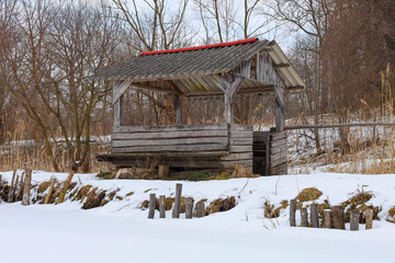 A wooden shelter stands on the bank of a snow-covered water body, surrounded by winter forest. Its grey slate roof is adorned with a red strip. The open front of the wooden structure faces the shore c © Олег Струс