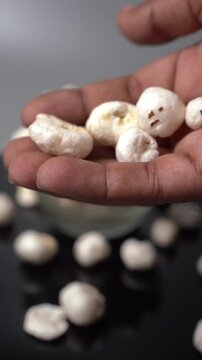 Fox nut or Euryale ferox snack presentation in a bowl with hand holding healthy puffed lotus seeds on a dark reflective surface