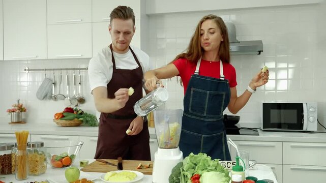 Young couple preparing healthy food together in a bright modern kitchen, with a woman pouring water into a blender while preparing fresh fruits and vegetables.
