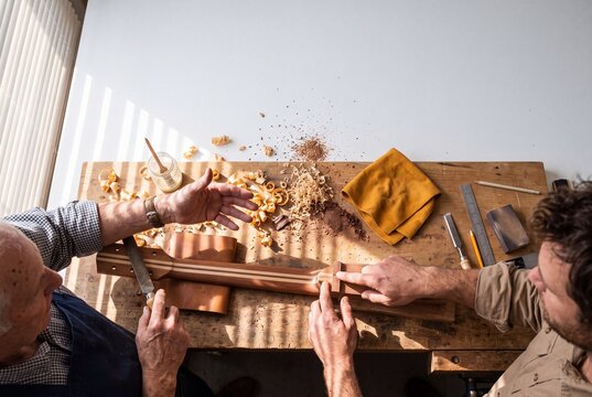 Senior and young luthiers inspect and shape a handcrafted wooden guitar neck at a workshop table covered with tools and wood shavings