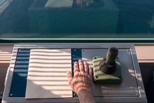 High-angle view of a captain's hand resting on a ship control panel with a joystick and paper by a window overlooking the ocean