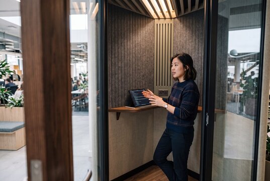 Young Asian businesswoman speaking on a video conference call inside an acoustic privacy booth in a modern office