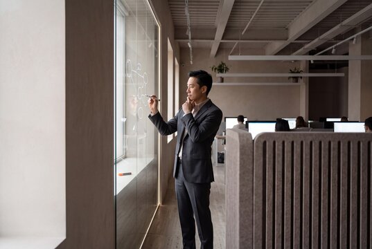 Asian businessman writing strategic workflow diagrams on a glass wall in a modern corporate office interior