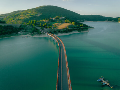Aerial view of Lago di Castreccioni with its long bridge crossing the turquoise water, surrounded by rolling hills and lush green landscape in Cingoli, Marche, Italy.