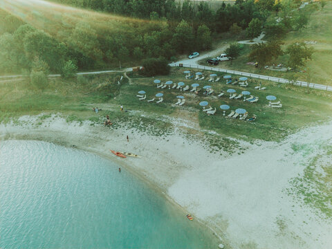 Aerial view of Lago Castreccioni with sun loungers, umbrellas, and kayaks on the sandy shore surrounded by lush green hills, Cingoli, Marche, Italy.