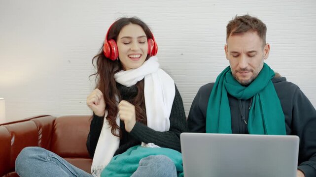 Young couple relaxing together at home during winter, with a woman enjoying music through headphones while a man works on a laptop.
