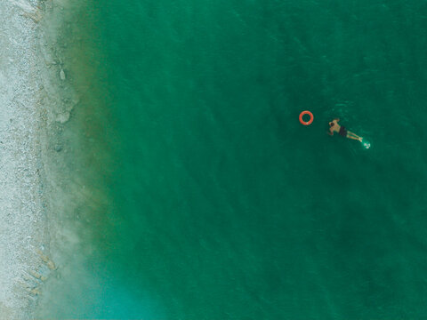 Aerial view of a person swimming with an orange lifebuoy in the turquoise waters of Lago Castreccioni near the pebbled shoreline in Cingoli, Marche, Italy.