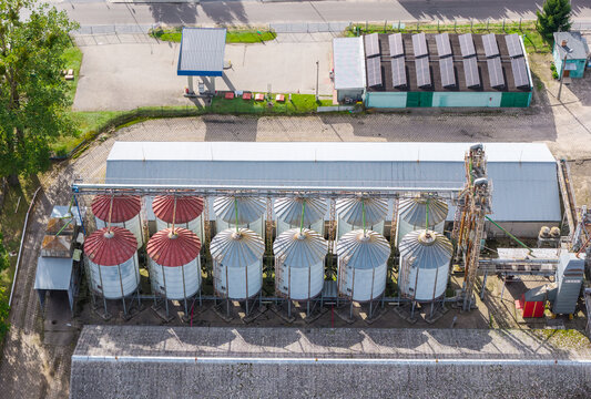 Aerial view of a grain elevator facility featuring multiple metal silos with red and silver roofs, connected by a conveyor system, situated next to industrial buildings with solar panels in Poland.