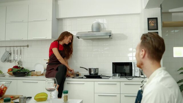 Woman preparing food on a stovetop while her partner watches from the foreground in a bright modern kitchen, with fresh ingredients nearby.
