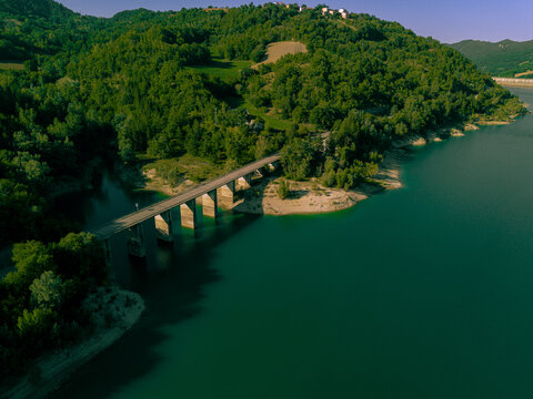 Aerial view of Lago di Gerosa with a bridge crossing the turquoise water surrounded by lush green hills and dense forests in Montemonaco, Marche, Italy.