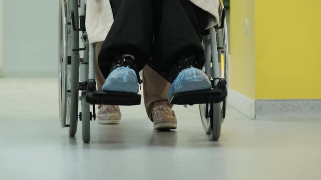 Nurse guides wheelchair toward sterile treatment office. Physician helps patient moving through bright medical corridor. Slow motion