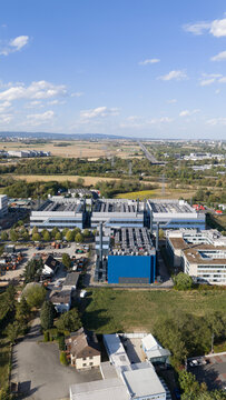Aerial view of a large industrial data center complex with blue and grey buildings surrounded by green fields and suburban infrastructure in Frankfurt am Main, Hessen, Germany.