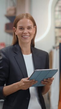 Vertical Screen. Clothing Store Shopping. Female Visual Merchandiser Holds a Tablet, Examining the Assortment on Display and Recording Stock Counts in the Digital System While Customers Browse