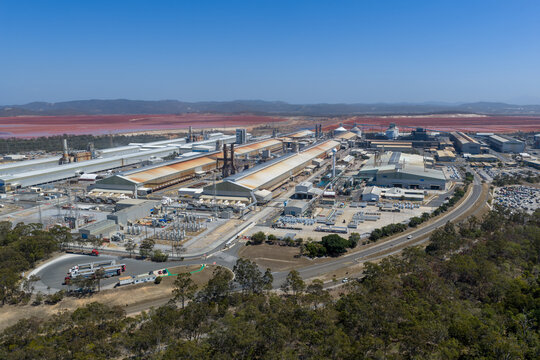 Aerial view of Boyne Smelter industrial facility with large factory buildings, storage tanks, and red tailings ponds under a clear blue sky in Boyne Island, Queensland, Australia.