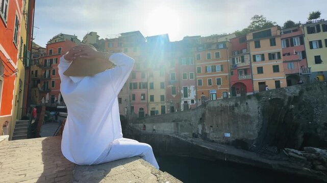 Fashion stylish woman sits on a wall overlooking the colorful houses of Riomaggiore, a picturesque village in Cinque Terre, Italy