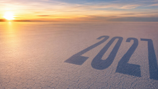 Large 2027 numerals cast a long shadow across cracked ground beside a calm horizon at sunset, suggesting a new year and future planning.