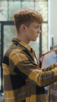 Vertical Screen. Portrait of a Stylish Young Man Shopping at Clothing Store, Selecting Menswear. Young Caucasian Boy Taking Items Off Hangers, Weighing Choices Before Buying in Retail Boutique