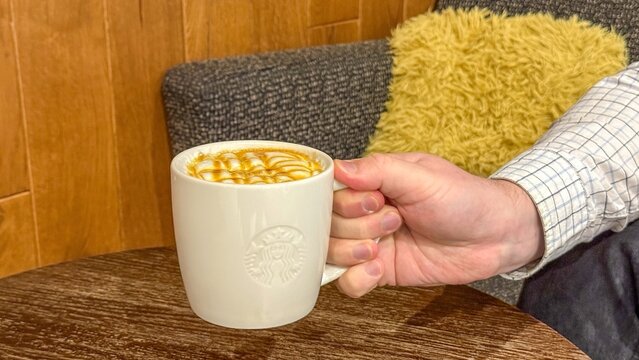 A person's hand holds a white  Starbucks mug filled with a beverage topped with foam, resting on a dark wooden table surface near a textured armchair 5 june 2026 Belgrade, Serbia