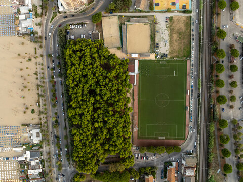 Aerial view of a football pitch adjacent to a dense pine forest and a sandy beach with rows of umbrellas in San Benedetto del Tronto, Marche, Italy.