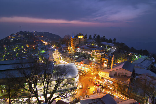 Aerial view of the illuminated town of Murree at dusk with snow-covered rooftops and mountain landscape in Murree, Punjab, Pakistan.