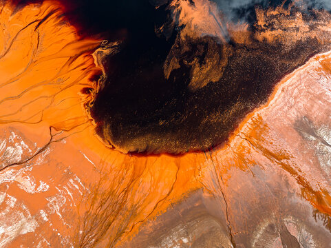 Aerial view of vibrant orange and red mineral-rich soil patterns surrounding a dark reflective pool in Pinjarra, Western Australia, Australia.