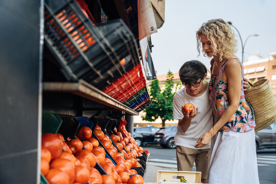 Teenage boy with Down syndrome inspecting ripe tomato beside his mother at outdoor market