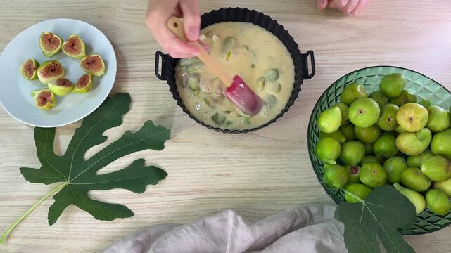 A person uses a red spatula to stir a creamy batter with pieces of green fruit in a black fluted pan, surrounded by whole figs and sliced figs on a table