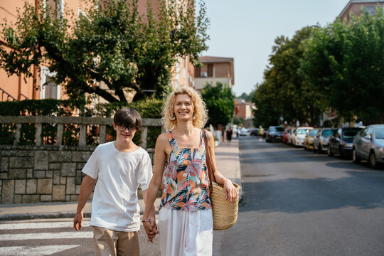Happy mature mother holding hands with teenage son with Down syndrome on sunny city street