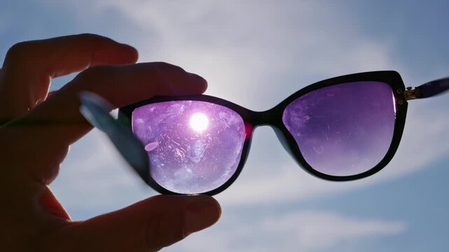 Hand holds dark sunglasses against bright sunlight outdoors. Vibrant purple lenses filter harsh glare under an open summer sky, providing protection and clear outdoor vision.
