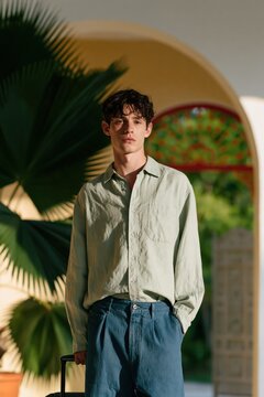 Young man in mint linen shirt and blue jeans standing by palm and arched doorway for summer fashion lookbook background