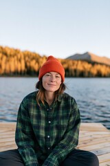 Fototapeta premium Young woman in orange beanie and plaid shirt on lakeside dock with autumn trees for outdoor portrait background