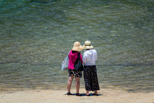 Two women in straw sun hats standing at the water's edge admiring the turquoise shallows of Hoshizuna beach, Yaeyama Islands, Iriomote, Japan