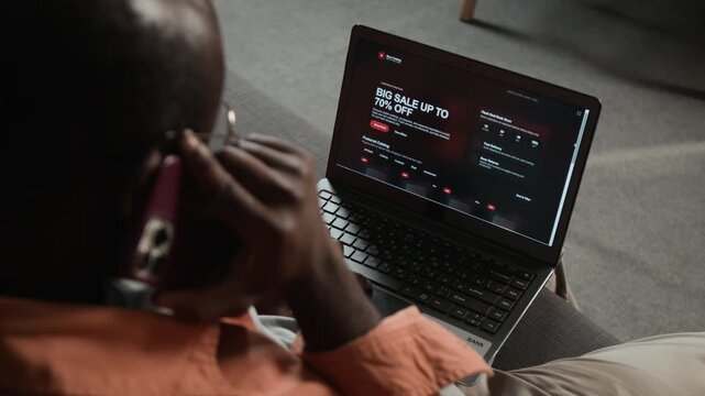 African American man browsing ecommerce store on laptop while talking on smartphone in over shoulder shot. Ideal for online shopping advertisements, consumer tech marketing, and retail campaigns