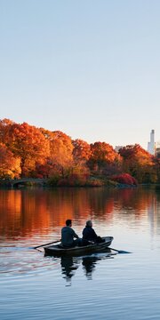 Two people rowing a small boat on a calm lake with orange autumn trees reflection for landscape background and peaceful nature banner