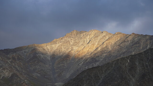 Golden-lit mountain peak at sunset with dramatic clouds in Jispa, India. Rugged terrain & warm golden hues make serene landscape in Lahaul Valley. high-altitude scenery & Himalayan range seen at HP.