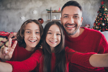 Naklejka premium Father and two children smiling for a Christmas family selfie in matching red sweaters by a decorated tree at home, celebrating holiday joy and togetherness