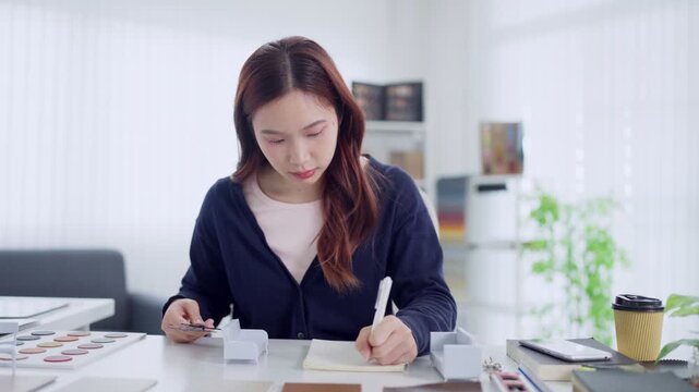 Young Asian furniture designer writing notes and reviewing material swatches at bright studio desk
