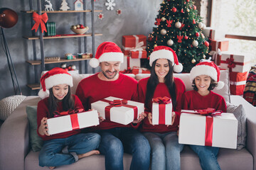 Naklejka premium Family opening Christmas gifts together on couch wearing Santa hats in cozy living room with decorated tree and wrapped presents