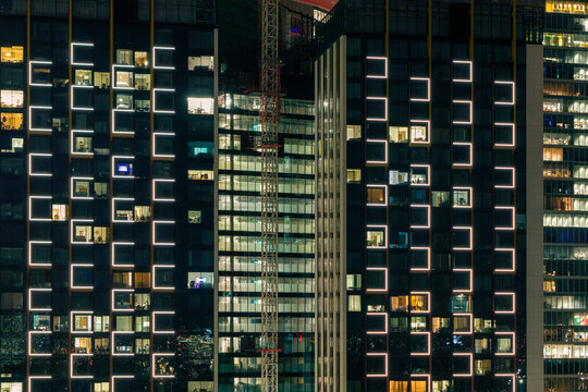 Aerial view of a modern office and residential building at night, featuring illuminated square window frames, glowing interior lights, and a prominent construction crane in Poland.