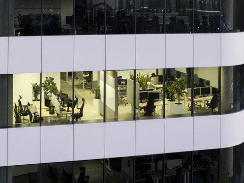 Aerial view of a modern office building with illuminated workspaces featuring desks, computers, and indoor plants visible through glass windows in Poland.