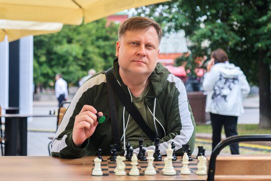 Adult man sitting at an outdoor chessboard in a park, deeply thinking and strategizing his next move in a classic intellectual board game.