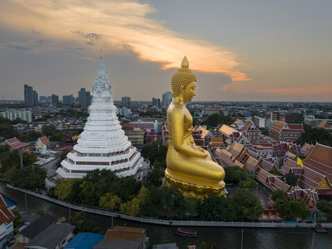 Aerial view of the golden Buddha statue and white stupa at Wat Paknam Phasi Charoen, surrounded by temple buildings and urban cityscape at sunset in Bangkok, Krung Thep Maha Nakho, Thailand.