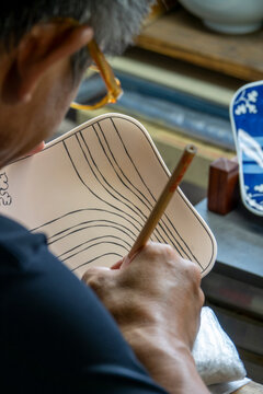 Bespectacled artisan drawing curved wave lines with a brush pen onto an unfired oval porcelain plate, Kyushu region, Arita, Japan