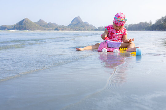 Asian Toddler Playing with Sand and Toy Truck at the Beach,An adorable young Asian toddler girl, wearing a patterned pink swimsuit and a matching patterned swim cap with goggles, happily plays in the 