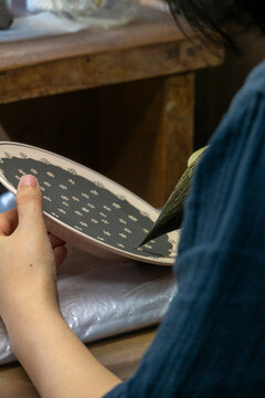 Woman's hand holding an oval plate while brushing black glaze around a white flower dot pattern, Kyushu region, Arita, Japan