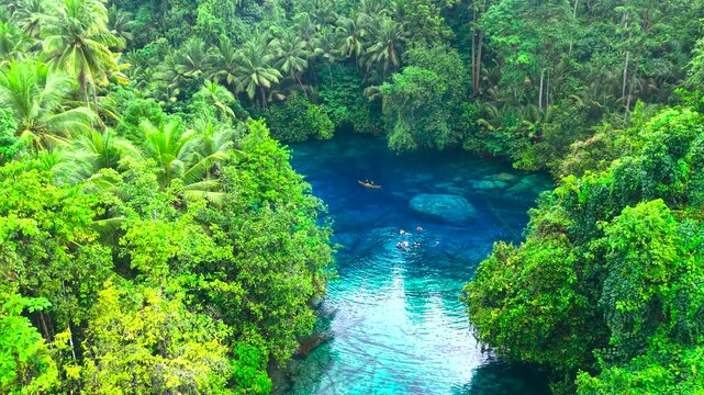 Aerial view of an adult man and woman paddling a canoe and adults swimming in a deep blue lake surrounded by tropical forest at Paisupok Lake, Indonesia.